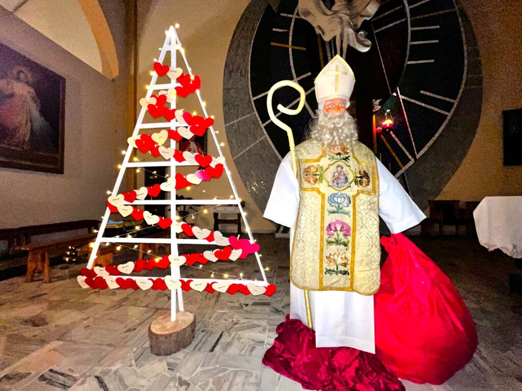 Rzeczywistej wielkości postać świętego Mikołaja ubranego w albę, ornat i mitrę, z pastorałem i workiem z prezentami w rękach. Obok sylwetka choinki na, której pozawieszane są papierowe serduszka