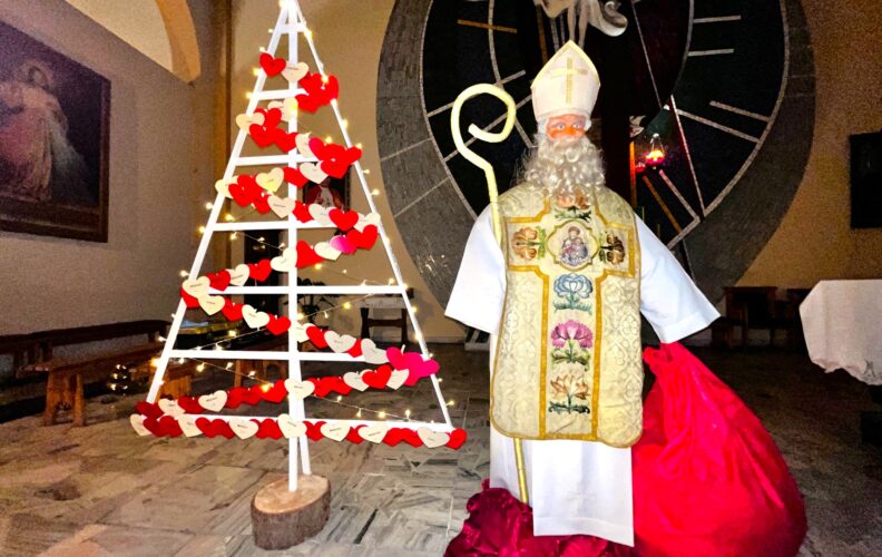 Rzeczywistej wielkości postać świętego Mikołaja ubranego w albę, ornat i mitrę, z pastorałem i workiem z prezentami w rękach. Obok sylwetka choinki na, której pozawieszane są papierowe serduszka